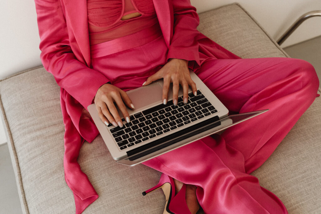 A woman dressed in a vibrant pink suit works on a laptop while seated on a beige couch. Her fingers rest on the keyboard, with matching pink heels visible beneath the laptop.